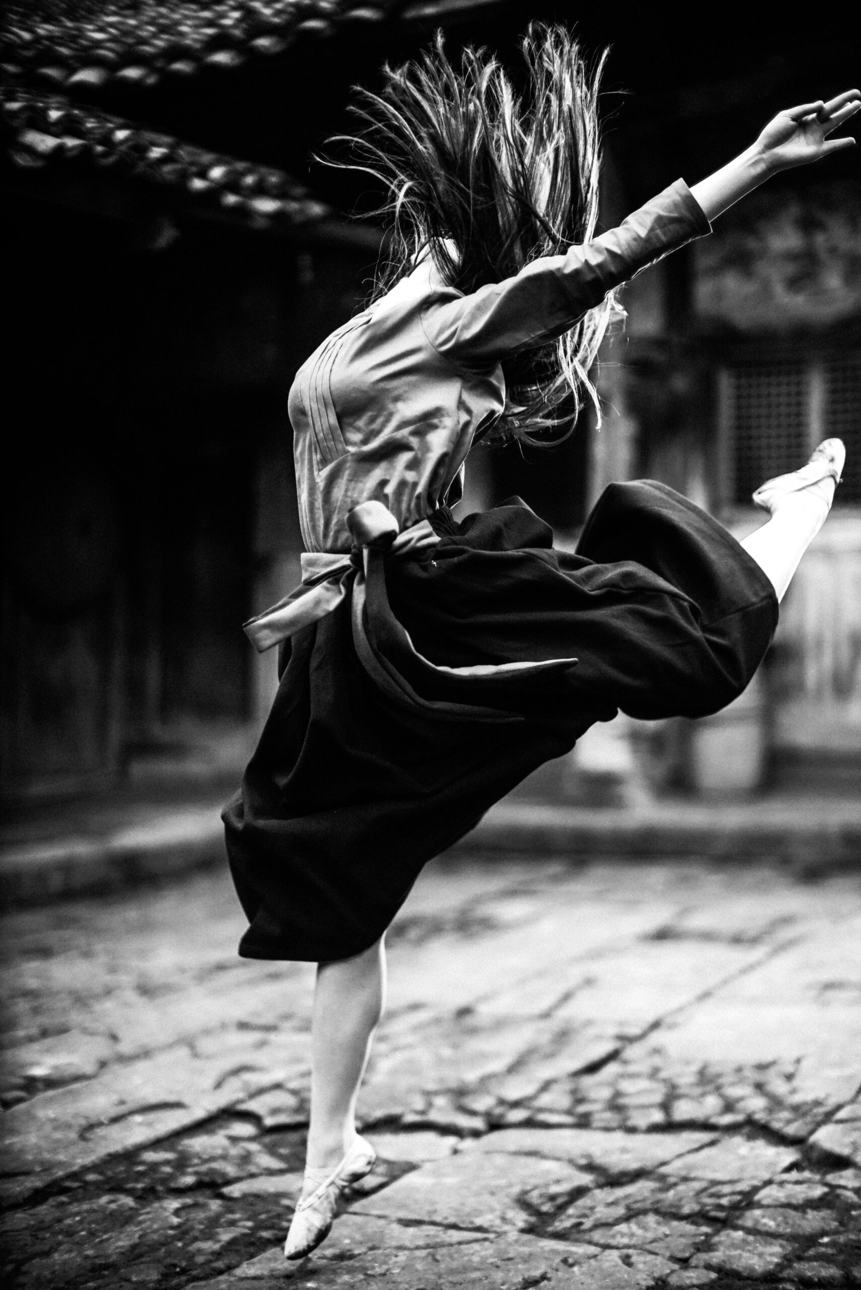 Black and white photo of a ballerina leaping gracefully on a cobblestone street.