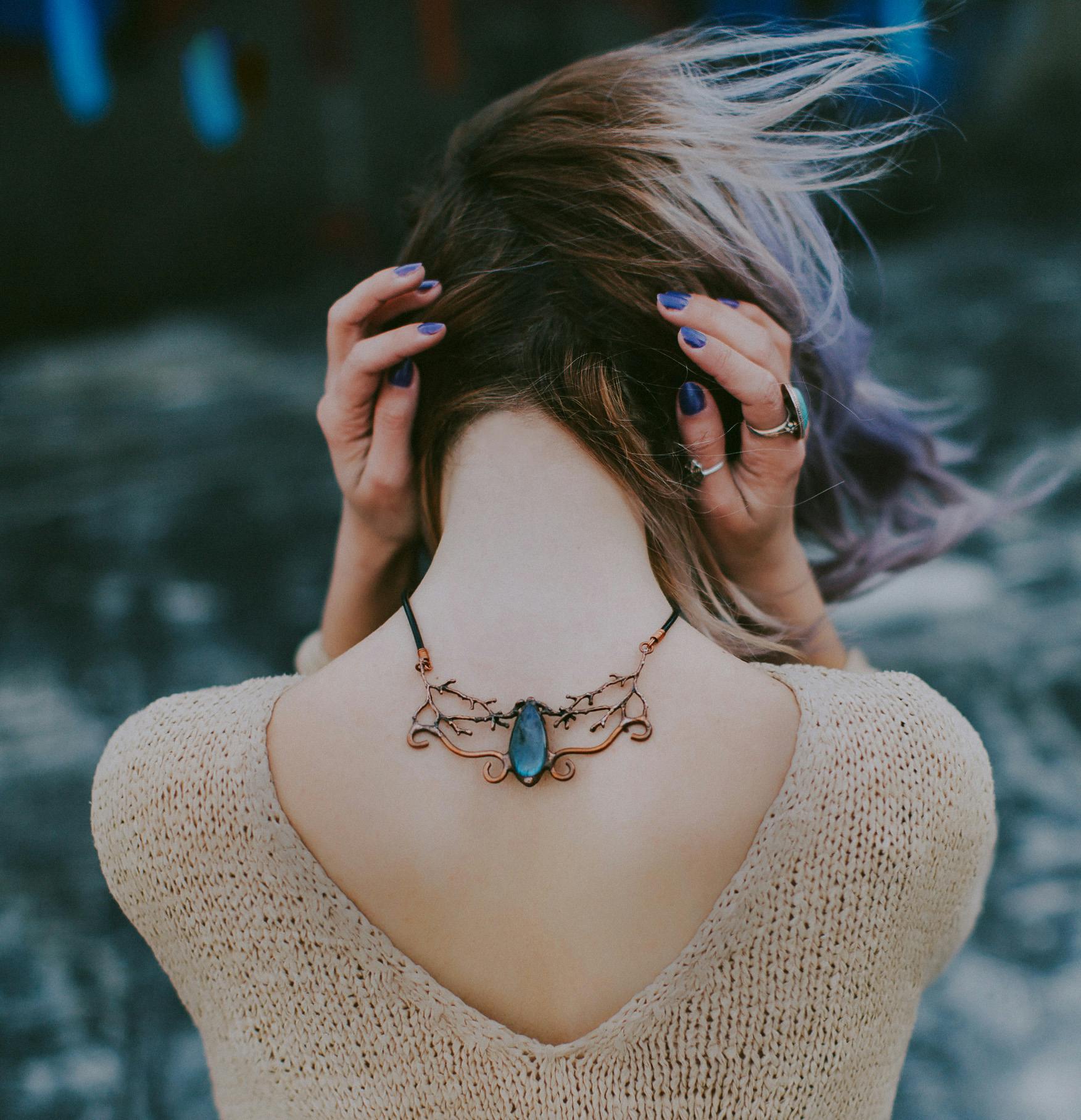 Glamorous young woman displaying a stylish gemstone necklace outdoors, posing with hands on head.
